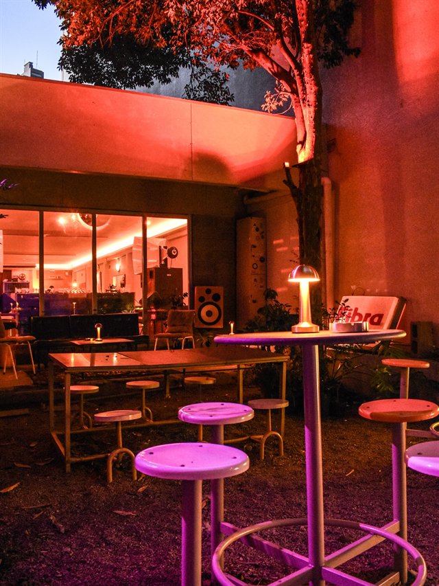 A casa vista de fora à noite, com a vidraça acesa em laranja e as mesas do pátio na frente.